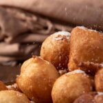 Brazilian sweet called "bolinho de Chuva", dumplings on a glass plate being sprinkled with sugar, on rustic wood, selective focus.